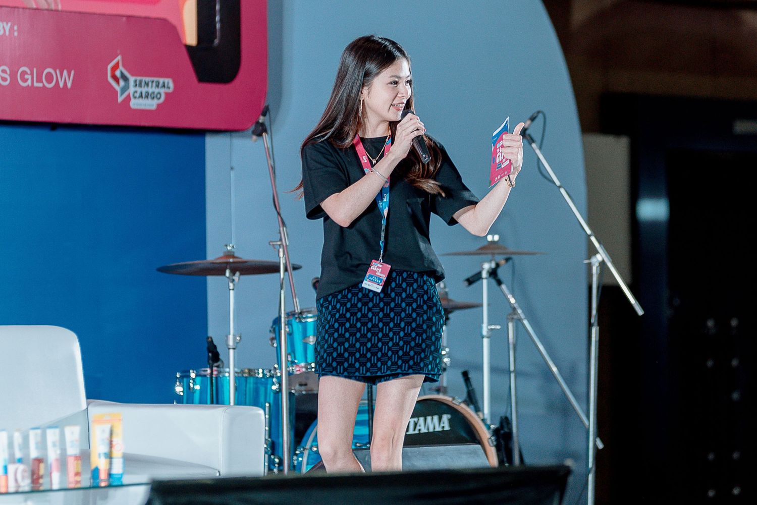 A performer in a polka dot dress sings on stage with a microphone stand and drum kit visible in the background.