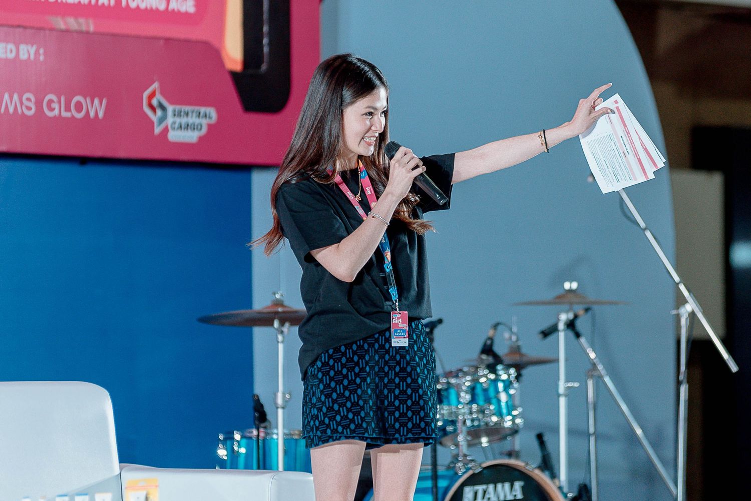 A performer in a black dress holds a microphone and papers while speaking on stage with a drum set in the background.