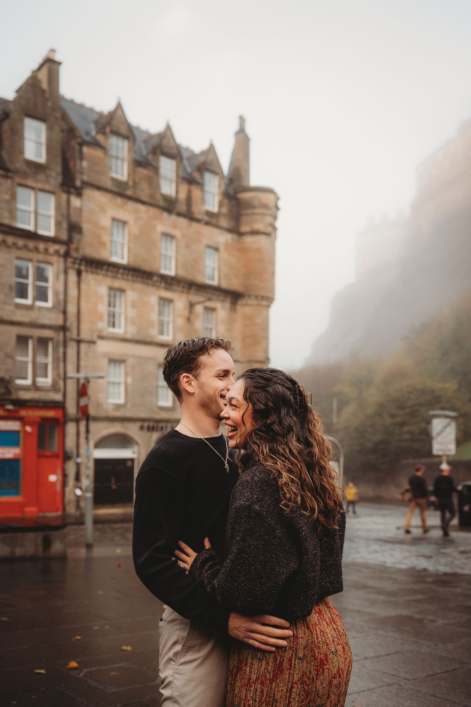 A couple embraces and laughs together on a misty street in front of historic Scottish stone buildings.