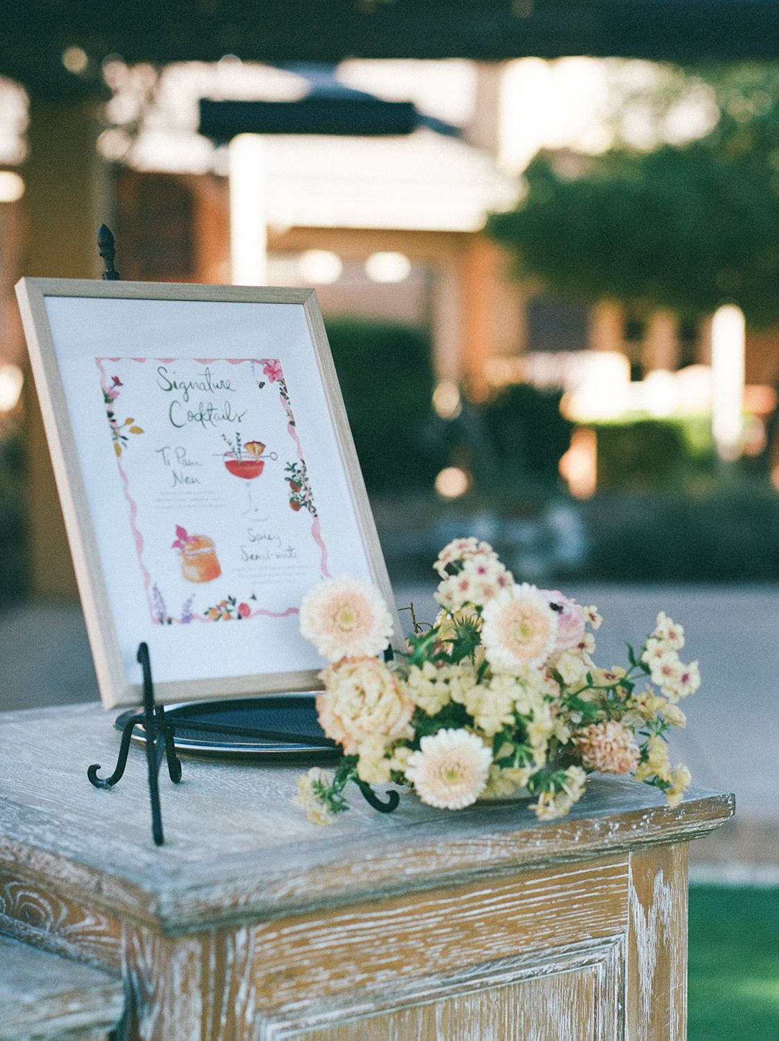 Pretty floral arrangement with white roses and a framed sign displayed on weathered rustic table at outdoor event setting.