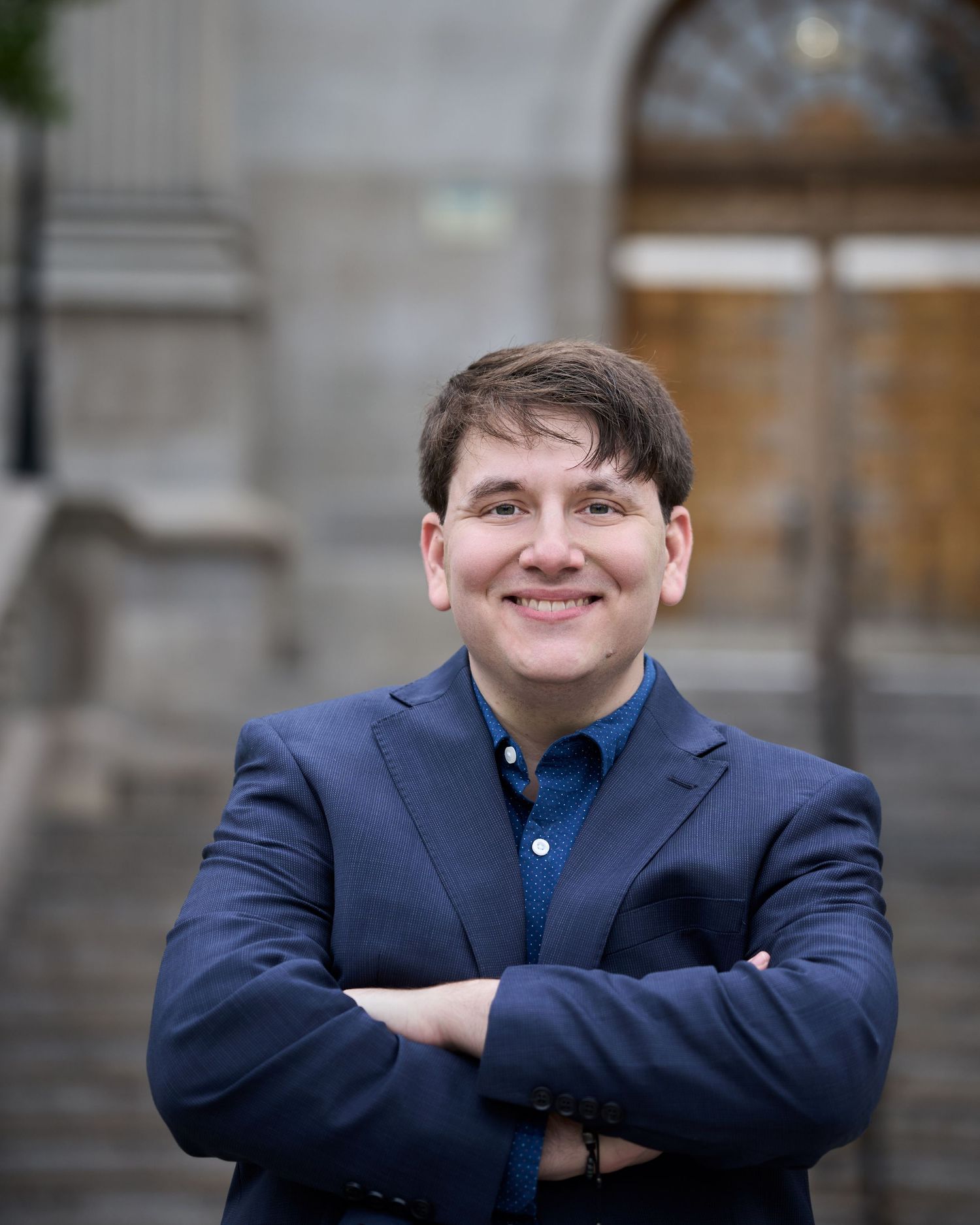 Professional headshot of someone in a navy blazer and blue button-up shirt standing with arms crossed outdoors.