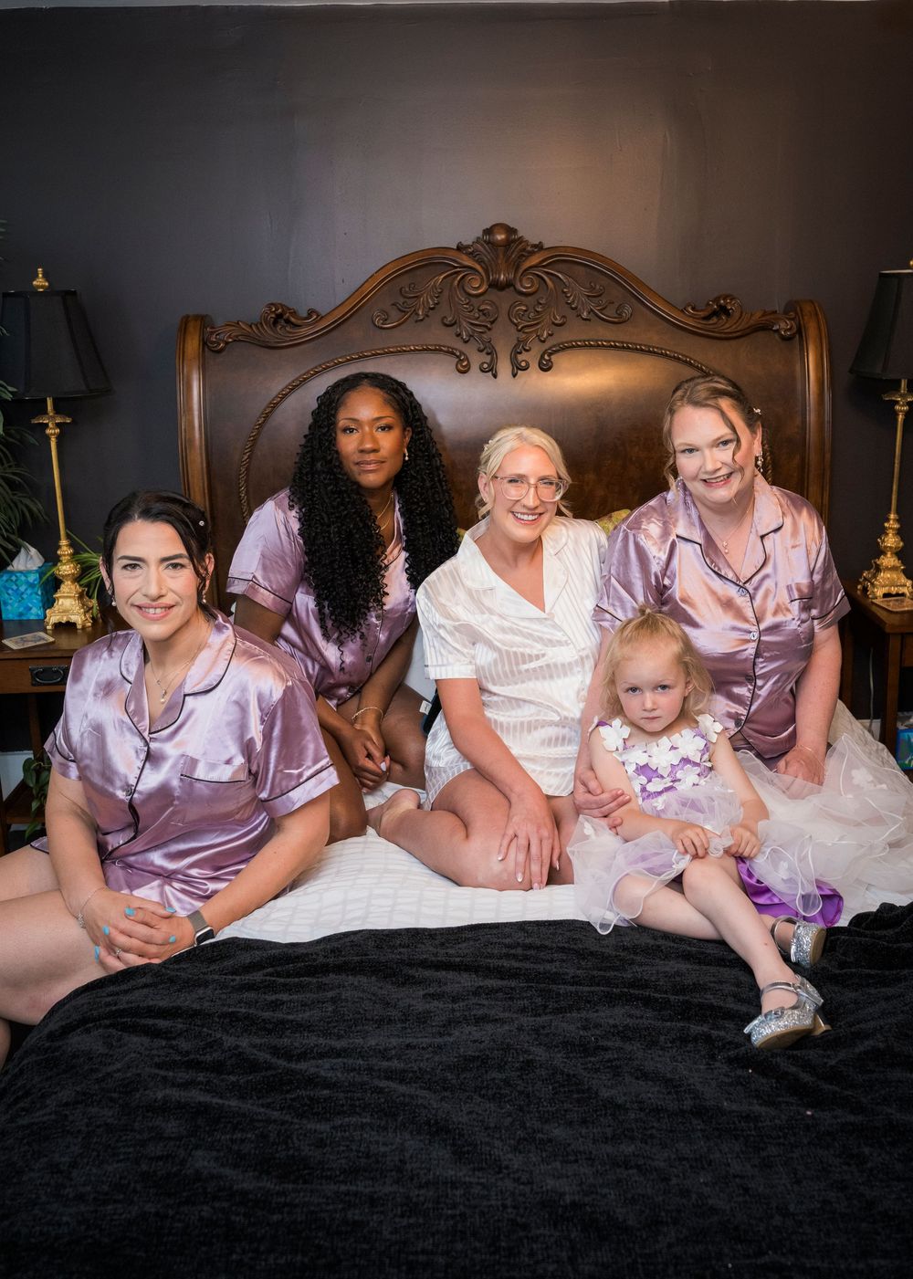 Bride poses with bridesmaids and flower girl in matching purple satin pajamas on an ornate bed.