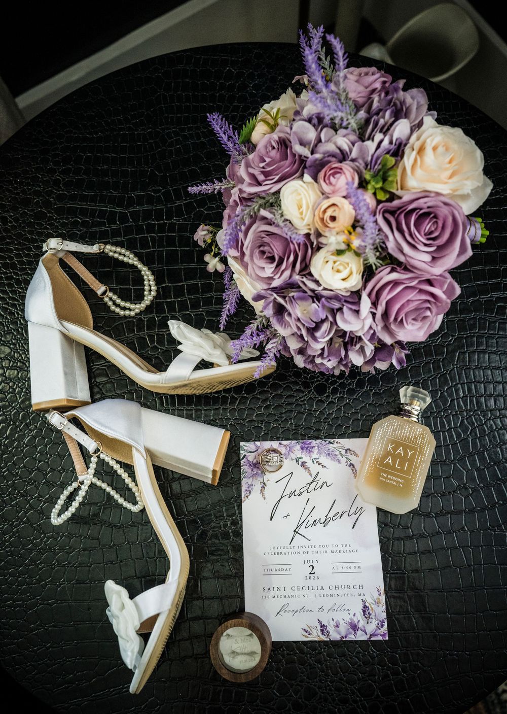 Wedding flat lay featuring purple rose bouquet, white heeled sandals, pearl bracelet, perfume, ring box, and floral invitation.