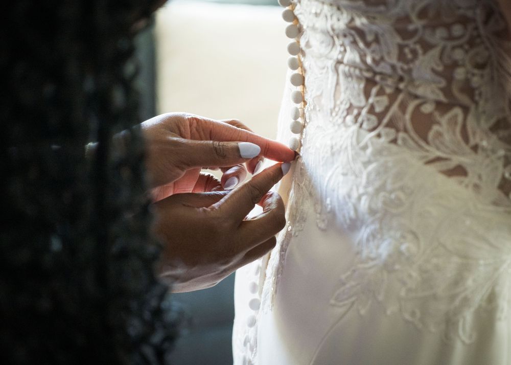 Close-up of hands fastening pearl buttons on an intricate lace wedding dress back.