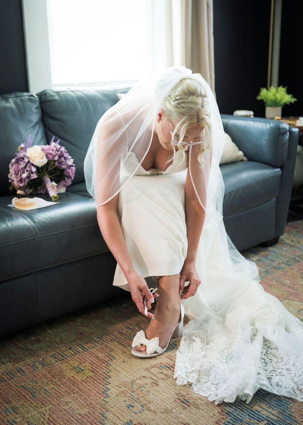 Bride in white wedding dress and veil adjusting her shoe while seated on a blue leather sofa.