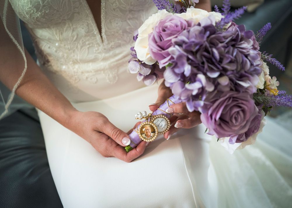 Bride holds a purple rose and hydrangea bouquet with a gold memorial photo charm on her wedding day.