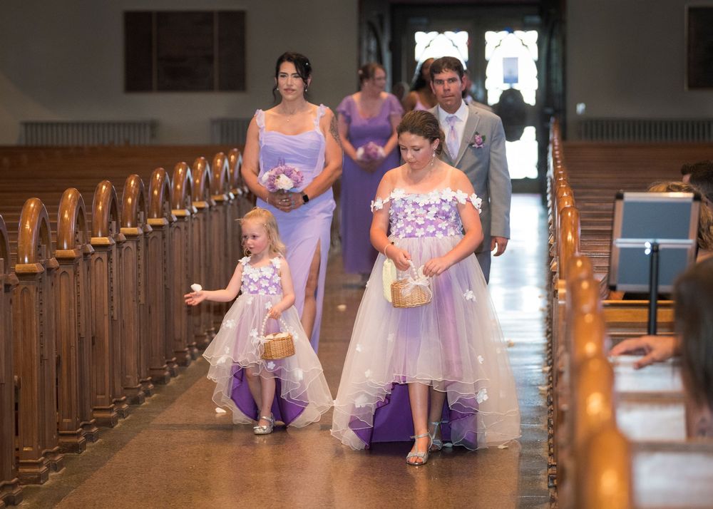 Bridesmaids and flower girls in lavender dresses walking down church aisle during wedding ceremony procession.