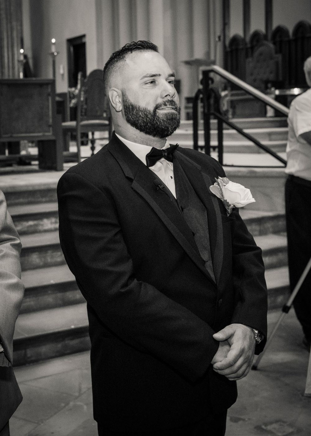 Black and white portrait of bearded groom in tuxedo with boutonniere watching bride walk down the aisle.