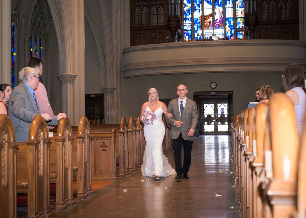 Bride walking down church aisle with escort past wooden pews and stained glass windows.