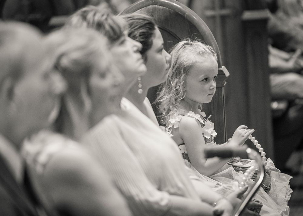Black and white photo of a young flower girl watching attentively during a wedding ceremony.