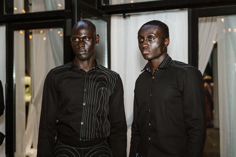 A group of people in black formal attire stand together at an indoor event with curtains and lighting in the background.