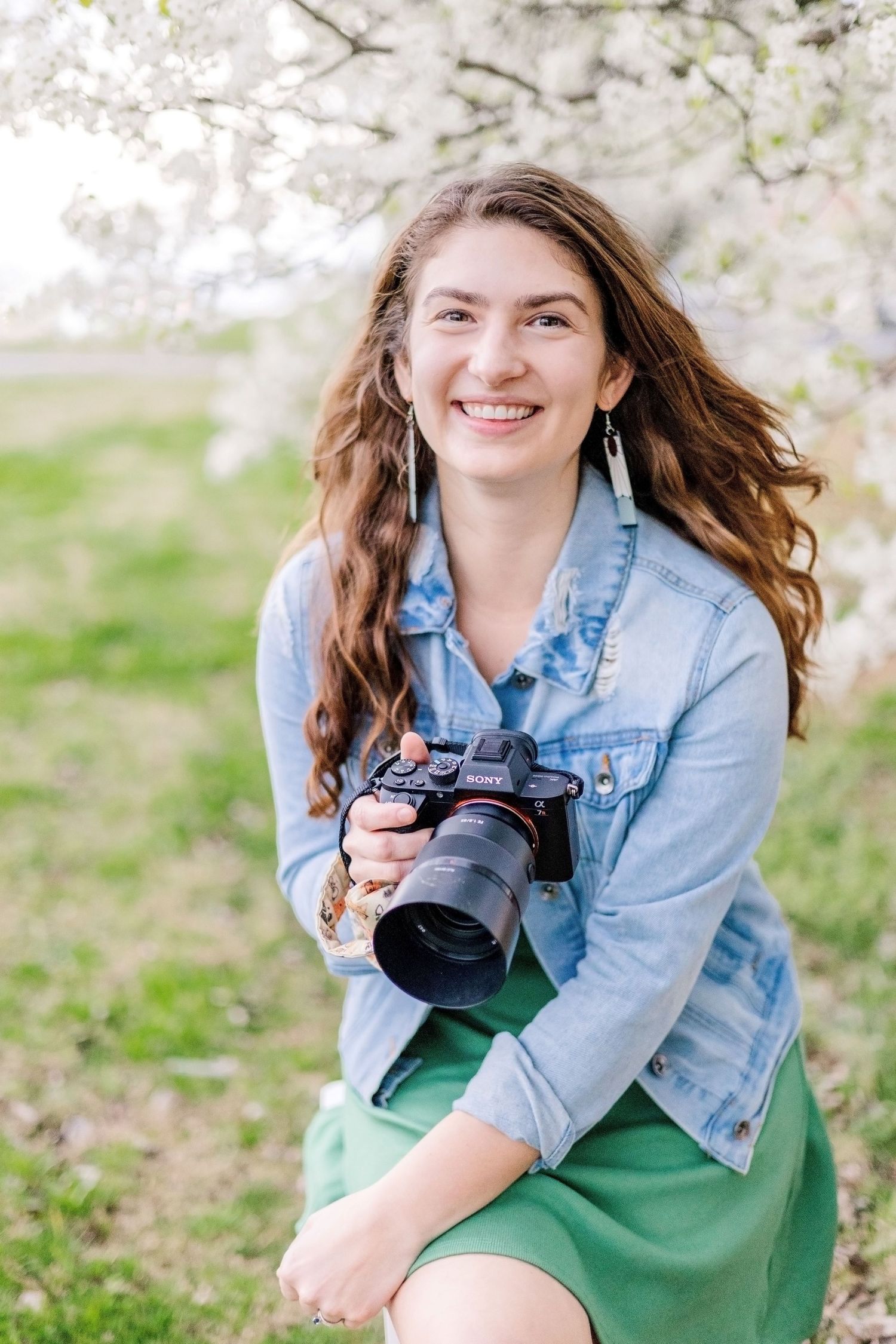 A photographer in a denim shirt holds a professional camera while standing outdoors with cherry blossoms in the background.