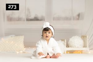 Small child wearing white hooded robe sits on white floor with decorative pillows and lights in background.