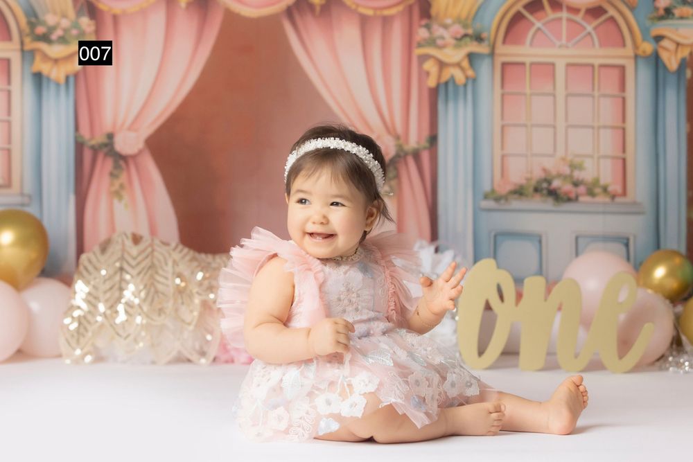 Smiling baby girl in pink tutu dress sits before colorful backdrop with balloons and \"one\" sign at first birthday.