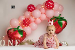 Baby girl in strawberry dress poses with pink balloon arch and strawberry foil balloons for a first birthday cake smash.