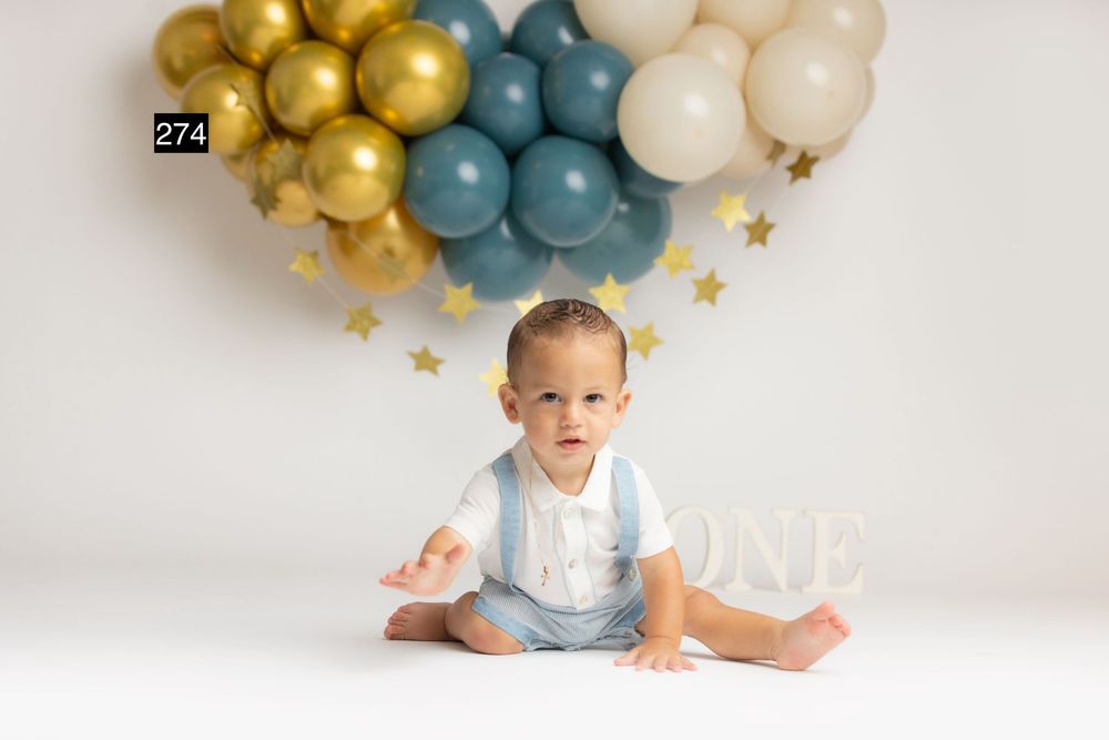 Baby boy in suspenders sits before gold and teal balloon heart display with ONE sign at first birthday cake smash.