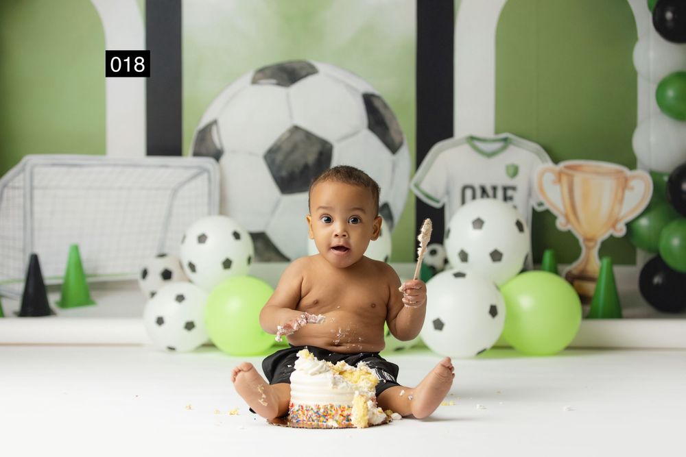 Baby boy sits smashing his first birthday cake surrounded by soccer-themed decorations, balloons, and a trophy backdrop.