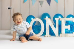 One year old baby boy sitting next to large blue and white ONE letters at first birthday photoshoot with bunting.