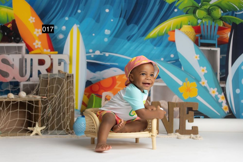 Baby in surf-themed photo shoot sitting on small bench with colorful beach backdrop, surfboards, and tropical decorations.