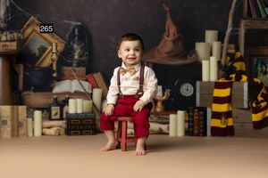 Toddler boy in suspenders and red pants sits on stool surrounded by Harry Potter themed books and candles.