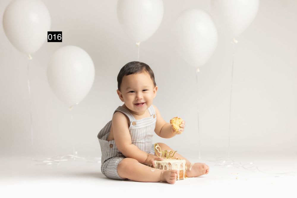 Smiling baby boy in gray romper sitting with white balloons and a gift box during a first birthday cake smash photo session.