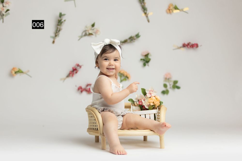 Smiling baby girl in white romper and bow headband sitting on small wooden bench with floral backdrop.