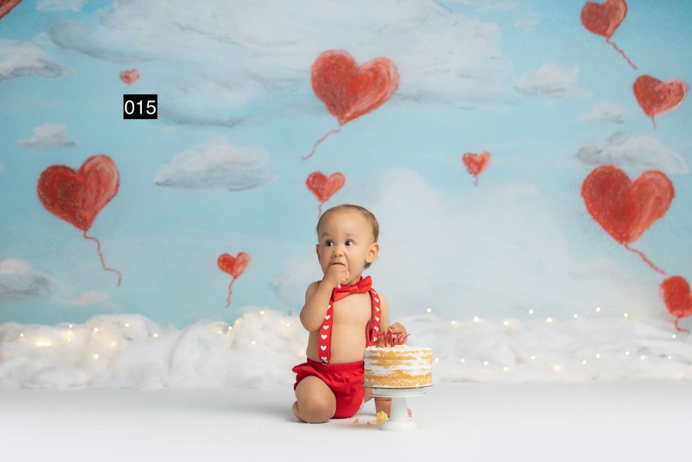 Baby in red romper sits by a small cake on a white floor with heart balloon backdrop for a first birthday cake smash.