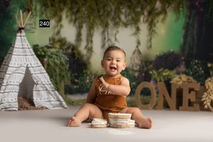 Laughing baby in brown romper sits beside a smash cake near a teepee with ONE letters for a first birthday photo.