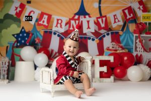 Baby in carnival outfit sits on white chair with colorful circus backdrop, balloons, and CARNIVAL banner for first birthday.