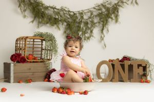 Baby girl in pink romper sits among strawberries beside wooden ONE letters for a first birthday cake smash photo.