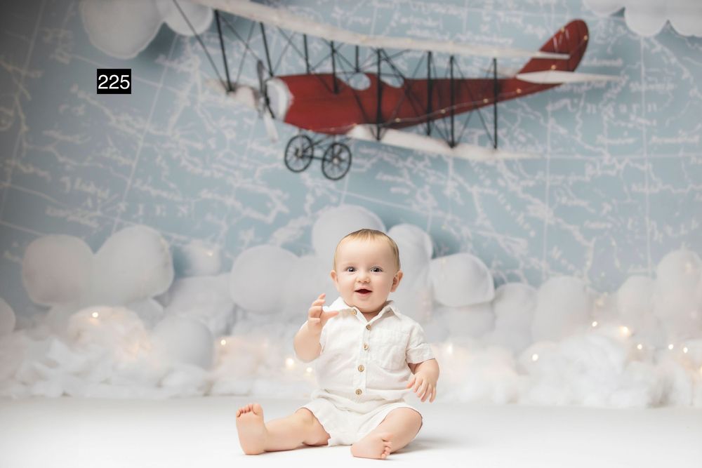 Baby in white outfit sits smiling before a whimsical backdrop featuring a red biplane soaring among fluffy clouds.