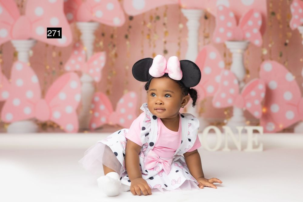 Baby girl in Minnie Mouse costume sits on white floor against pink polka dot backdrop celebrating first birthday.