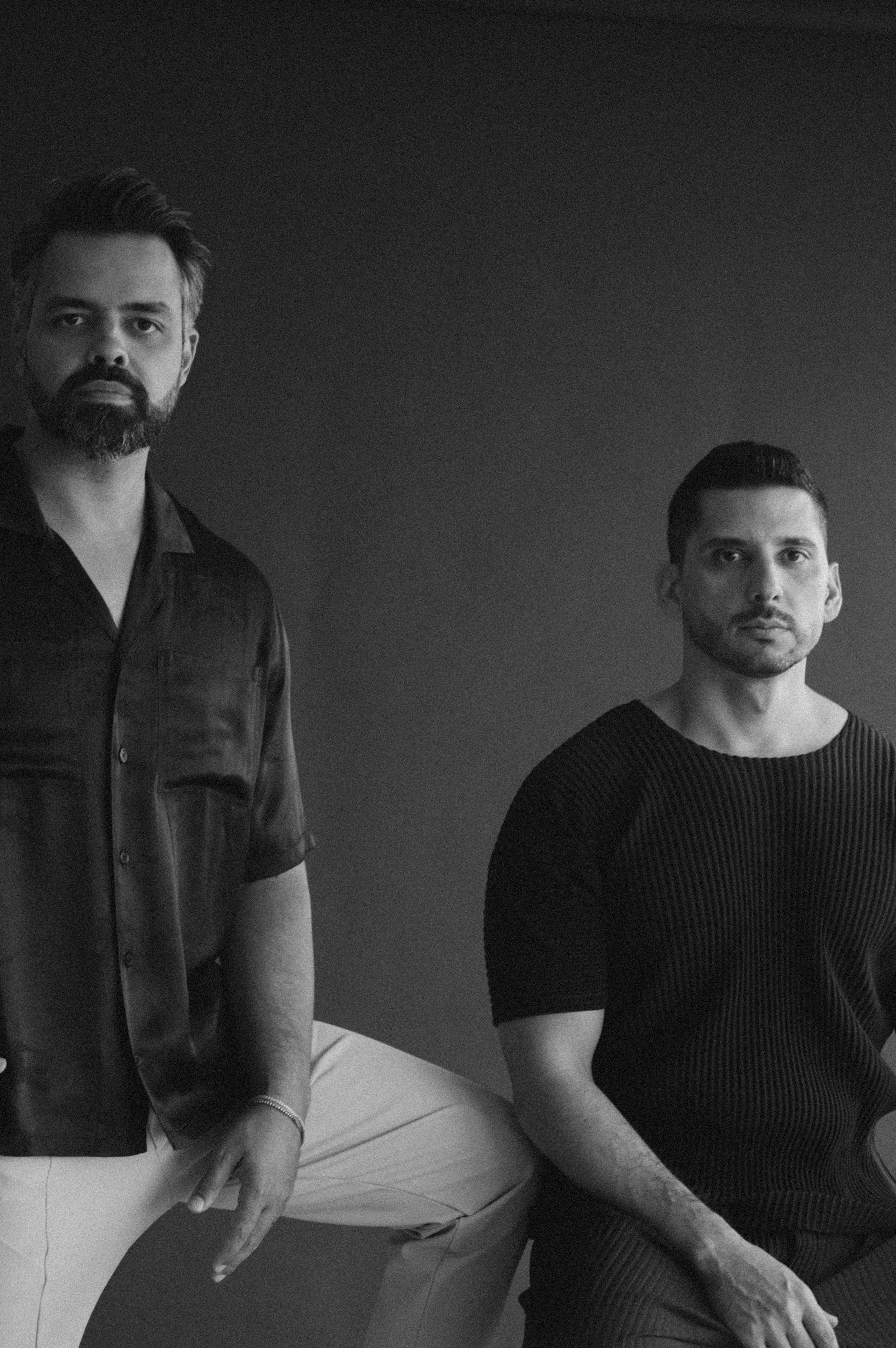 Professional studio portrait of two men in casual attire against a dark background.