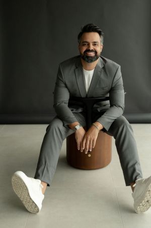 Portrait of a man in a grey suit and white sneakers sitting on a brown cylindrical ottoman.