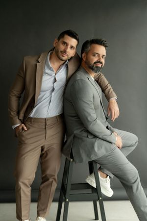 Fashionable studio portrait of two men in suits posing against a dark grey background.