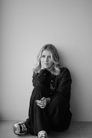 Black and white photo of someone in dark clothing sitting on floor against wall holding a phone.