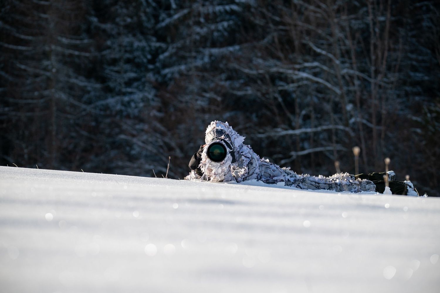 A sniper in winter camouflage lies prone in deep snow with a rifle, aiming into a dark forest background.