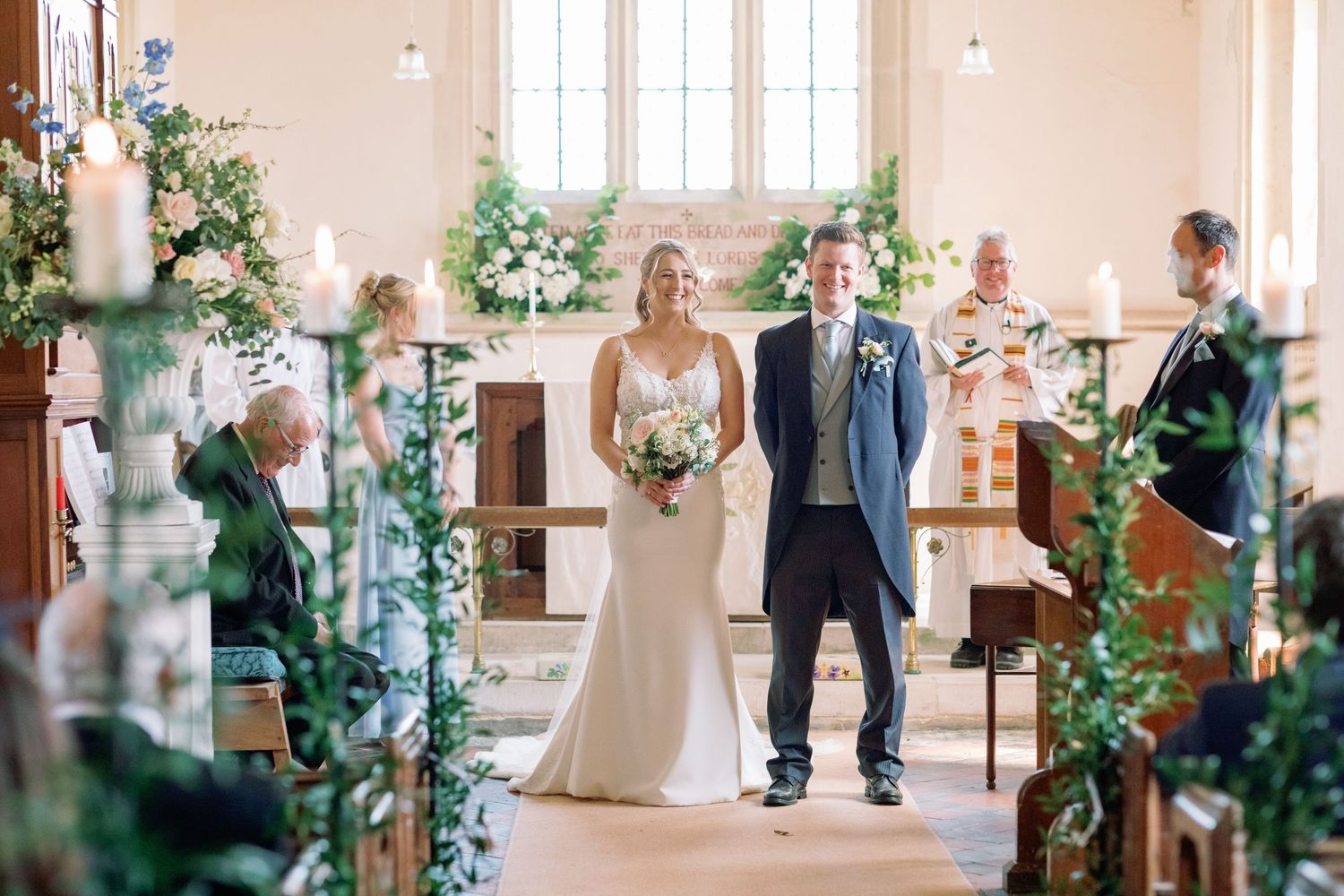 A couple stands at the altar during their wedding ceremony in a church decorated with greenery and candles.