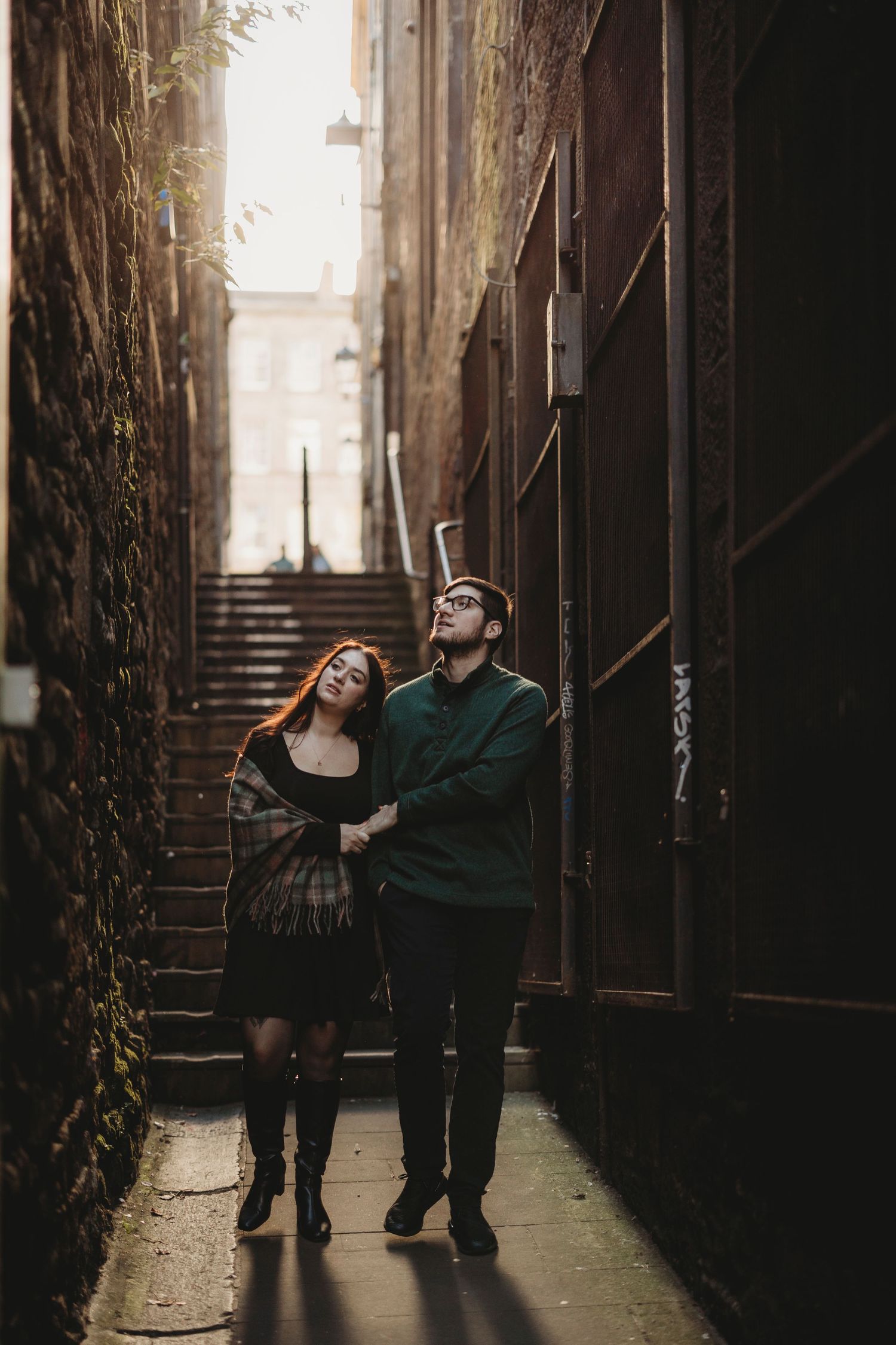 A couple dances playfully in a dimly lit urban alley with dramatic lighting and ivy-covered walls.