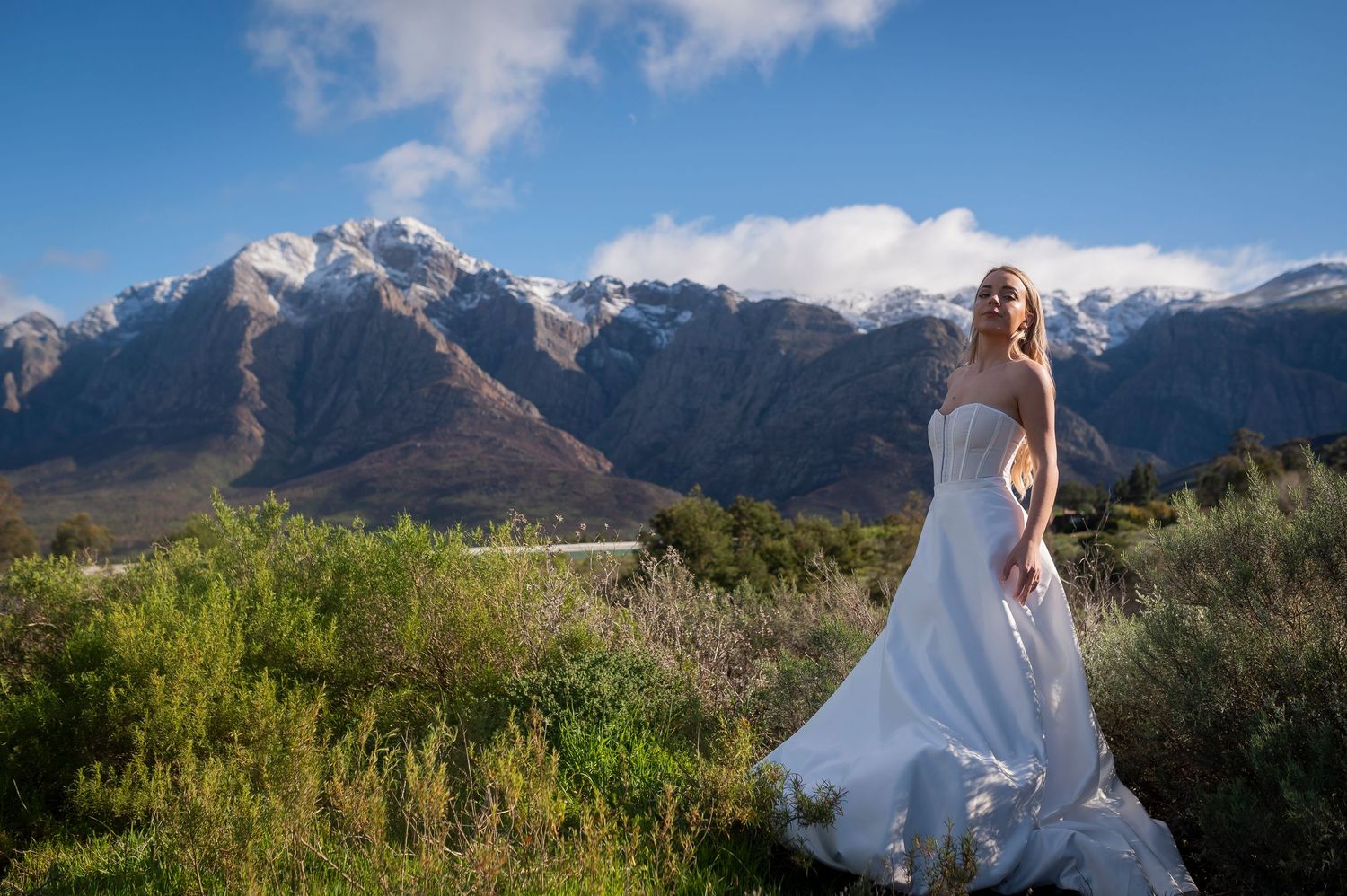 Bride in white gown poses before snow-capped mountains and lush greenery under a bright blue sky.