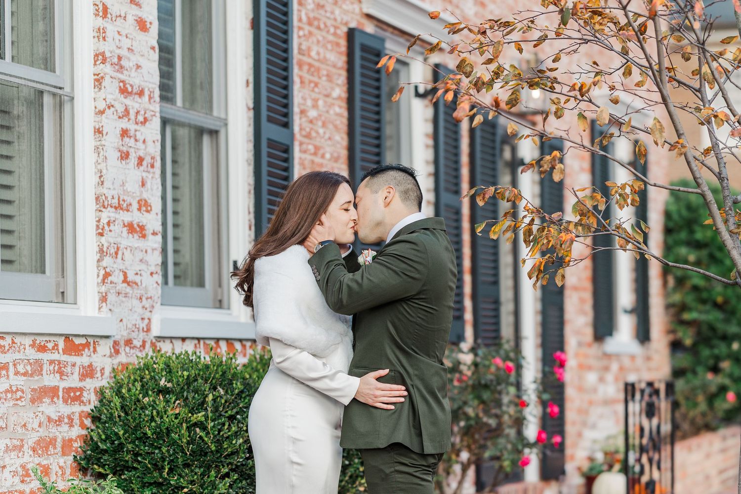 A couple stands in front of a brick house with white trim and autumn leaves falling around them.