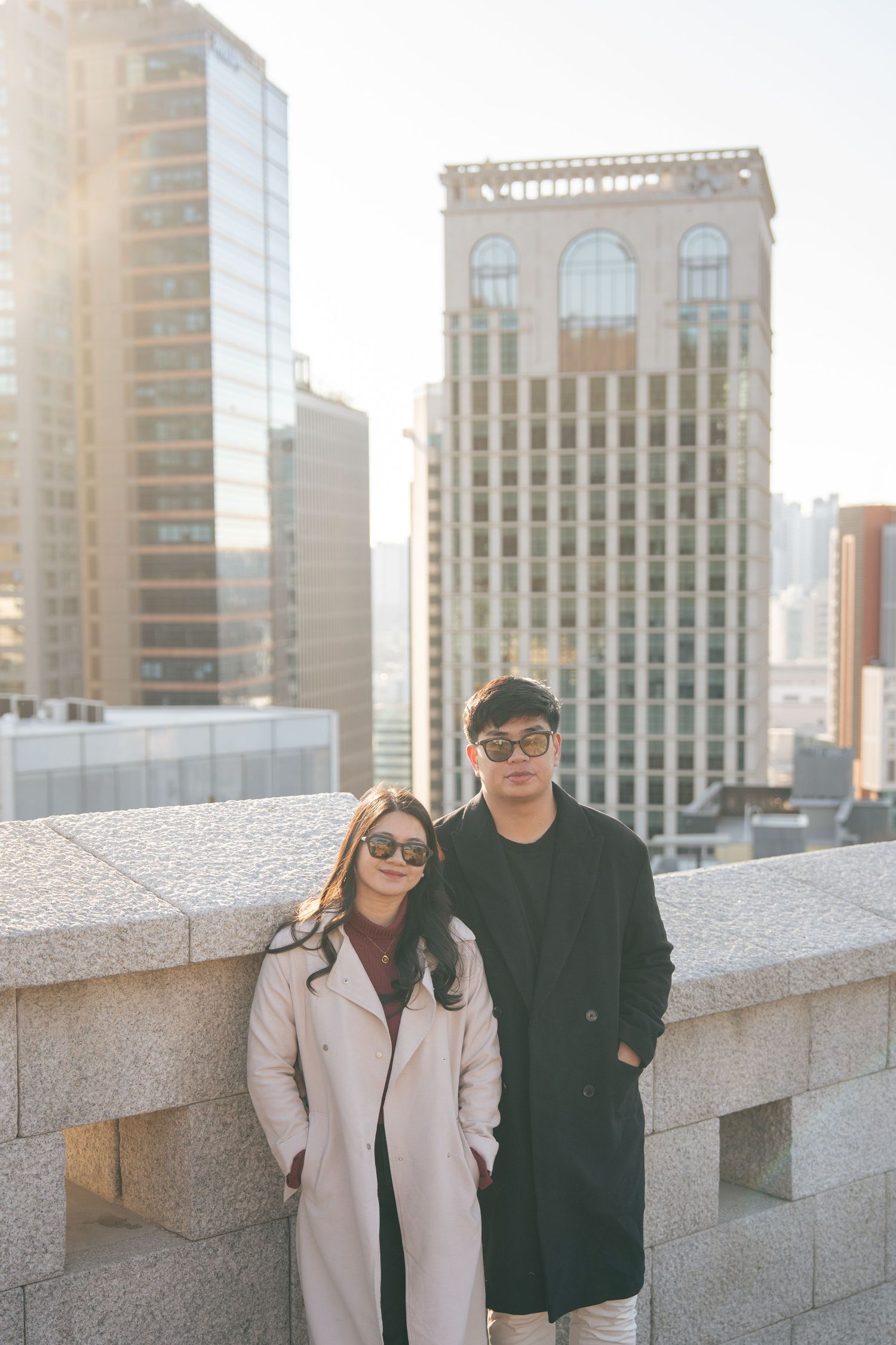 Two people in long coats pose together on an urban rooftop with city buildings and sunlight in the background.