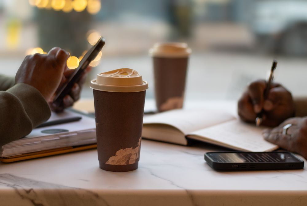 Coffee cups and notebooks on table with person using mobile phone.