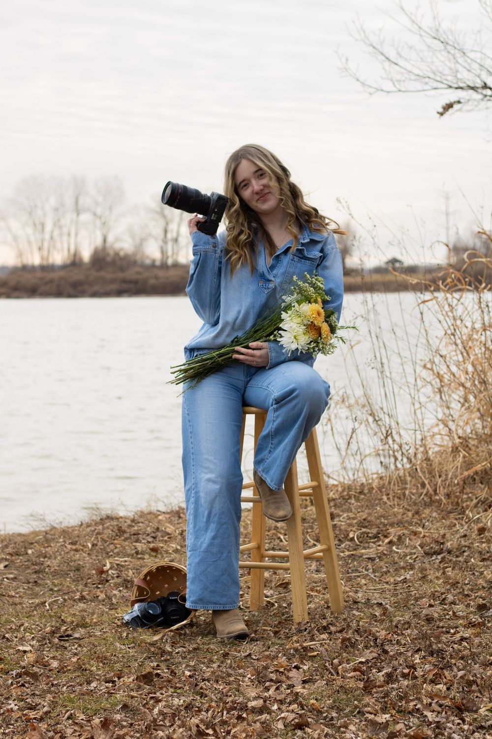 Photographer in denim outfit sits on wooden stool by lake holding camera and sunflower bouquet.