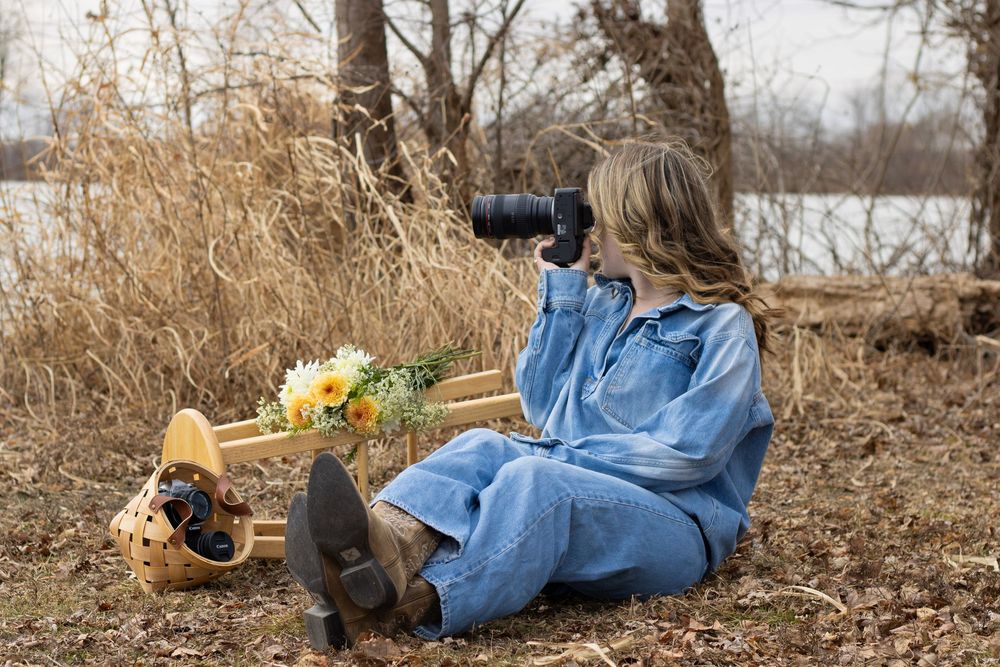 Photographer in denim relaxes on ground with camera and flowers by lake.