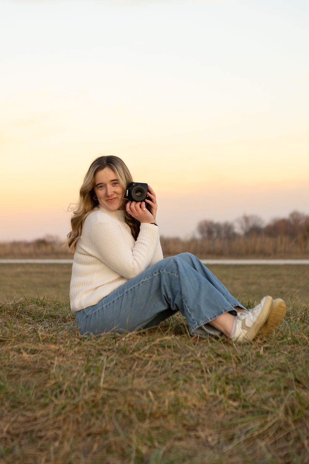 Photographer in cream sweater and jeans sits on grass during golden hour with camera.