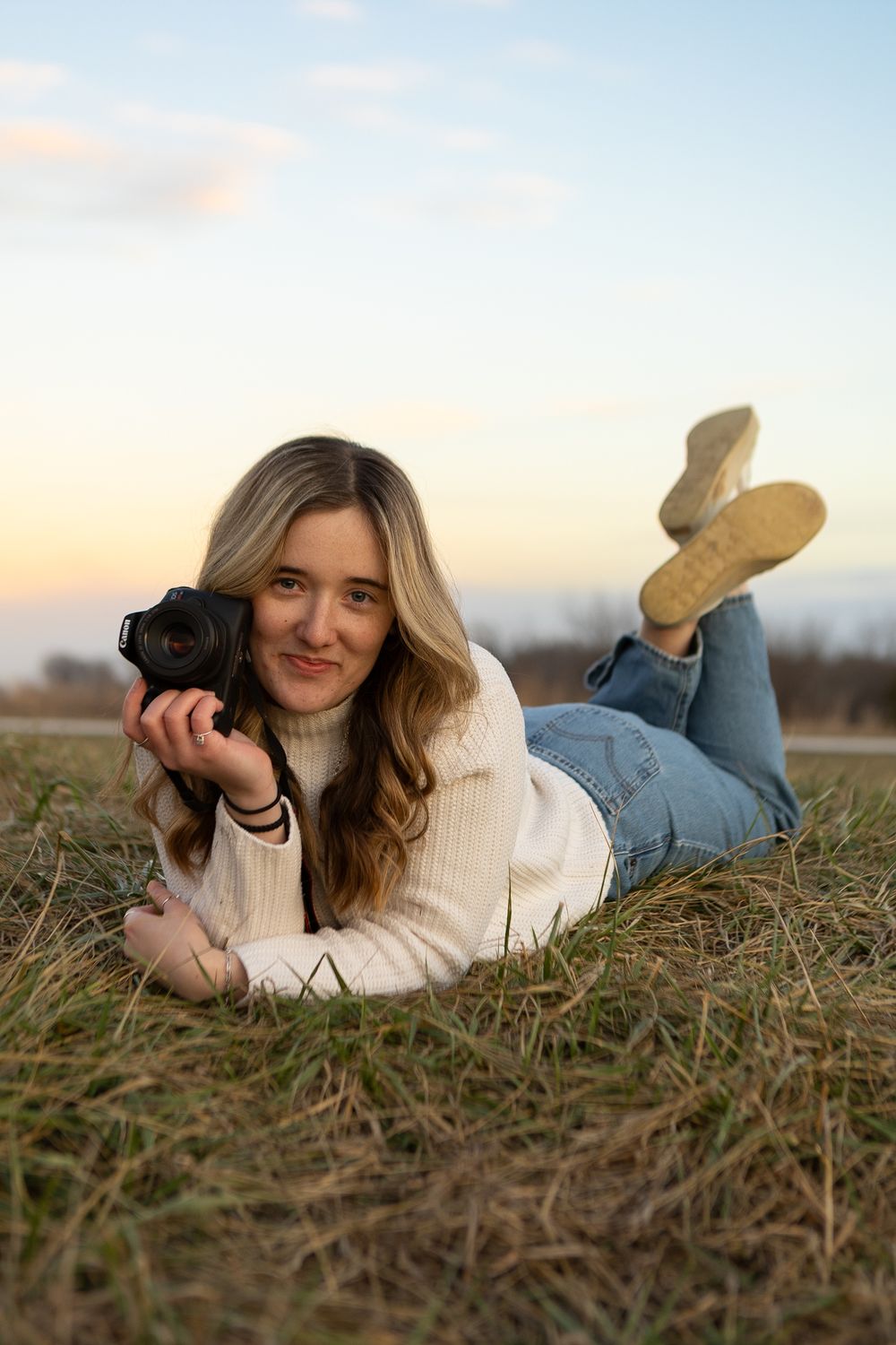 Photographer in cozy sweater lies on grass with camera during sunset.