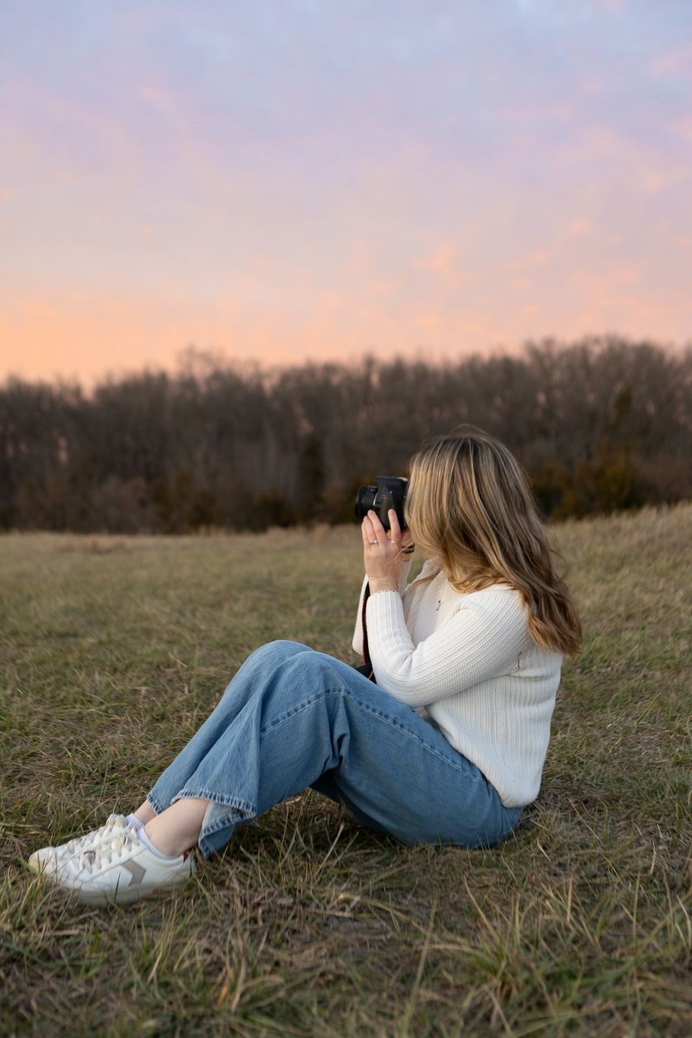 Photographer in white sweater takes photos while sitting in field at sunset.