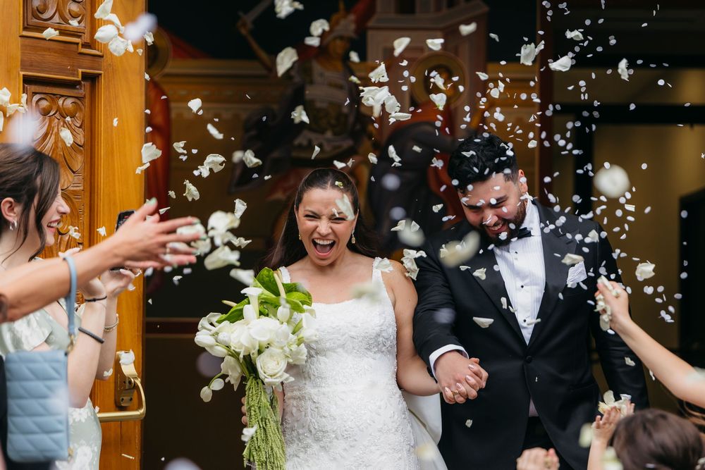 A joyful wedding couple exits their ceremony while guests shower them with white confetti and flower petals.