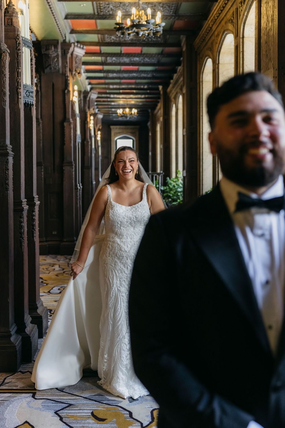 A bride in a lace wedding dress stands in a hallway with columns while a groom in a tuxedo is in the foreground waiting for fist look
